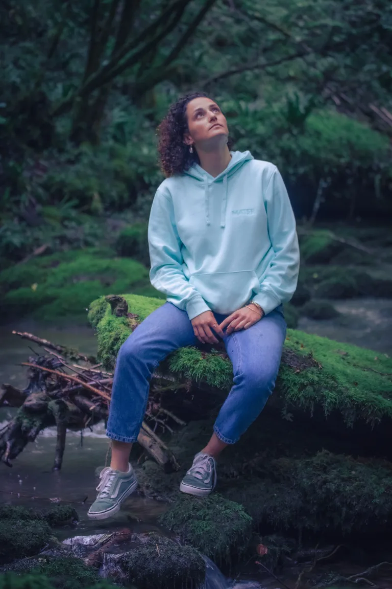 Une femme assise sur un rocher les pieds au dessus de l’eau au bon milieu de la rivière regarde le ciel pensive, elle porte le sweat-shirt de l’eau de la collection Lunélys Création