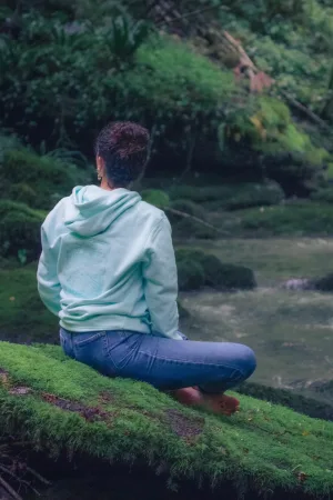 Photo d'une femme en tailleur sur un rocher moussu en pleine méditation regarde de rpofil dos à la caméra portant le sweat de l'eau de la nouvelle collection de Lunélys Création