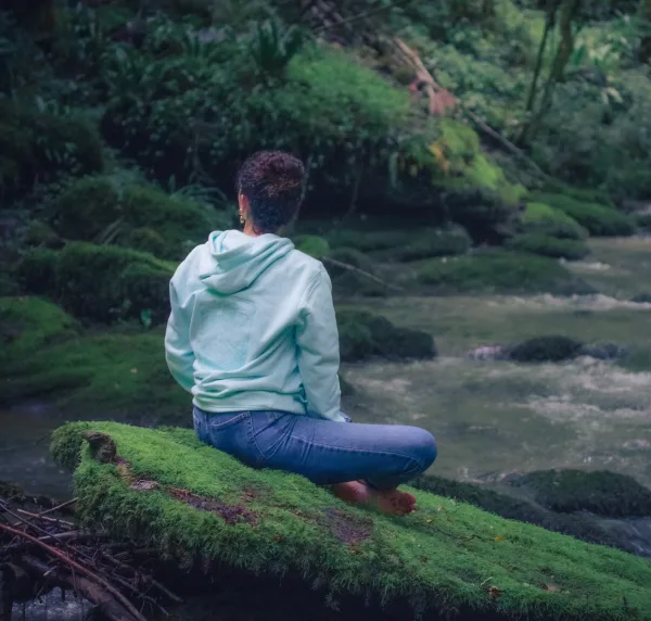 Photo d'une femme en tailleur sur un rocher moussu en pleine méditation regarde de rpofil dos à la caméra portant le sweat de l'eau de la nouvelle collection de Lunélys Création