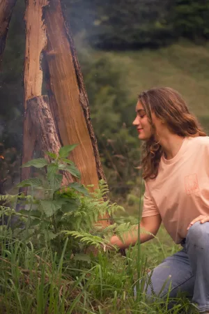 Femme portant un t-shirt beige, accroupie près d’un feu de camp entouré de bûches et de verdure.