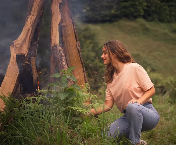 Femme portant un t-shirt beige, accroupie près d’un feu de camp entouré de bûches et de verdure.
