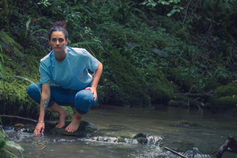 Photo d'une femme accroupis touchant l'eau dans la forêt, en fixant la caméra et portant le t-shirt de la collection WATER de Lunélys Création